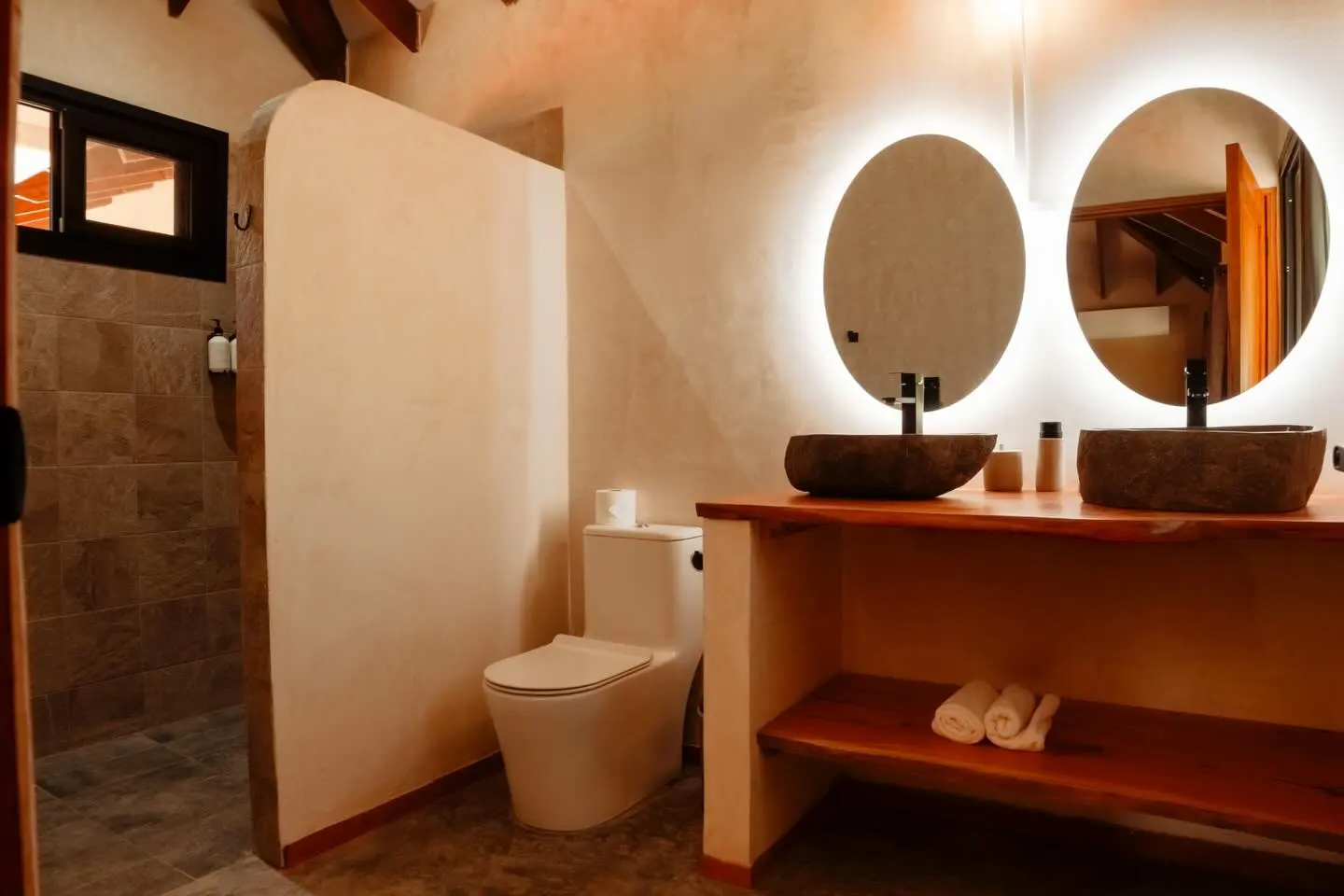 Baño con dos lavamanos de piedra sobre una repisa de madera y dos espejos redondos con luz.