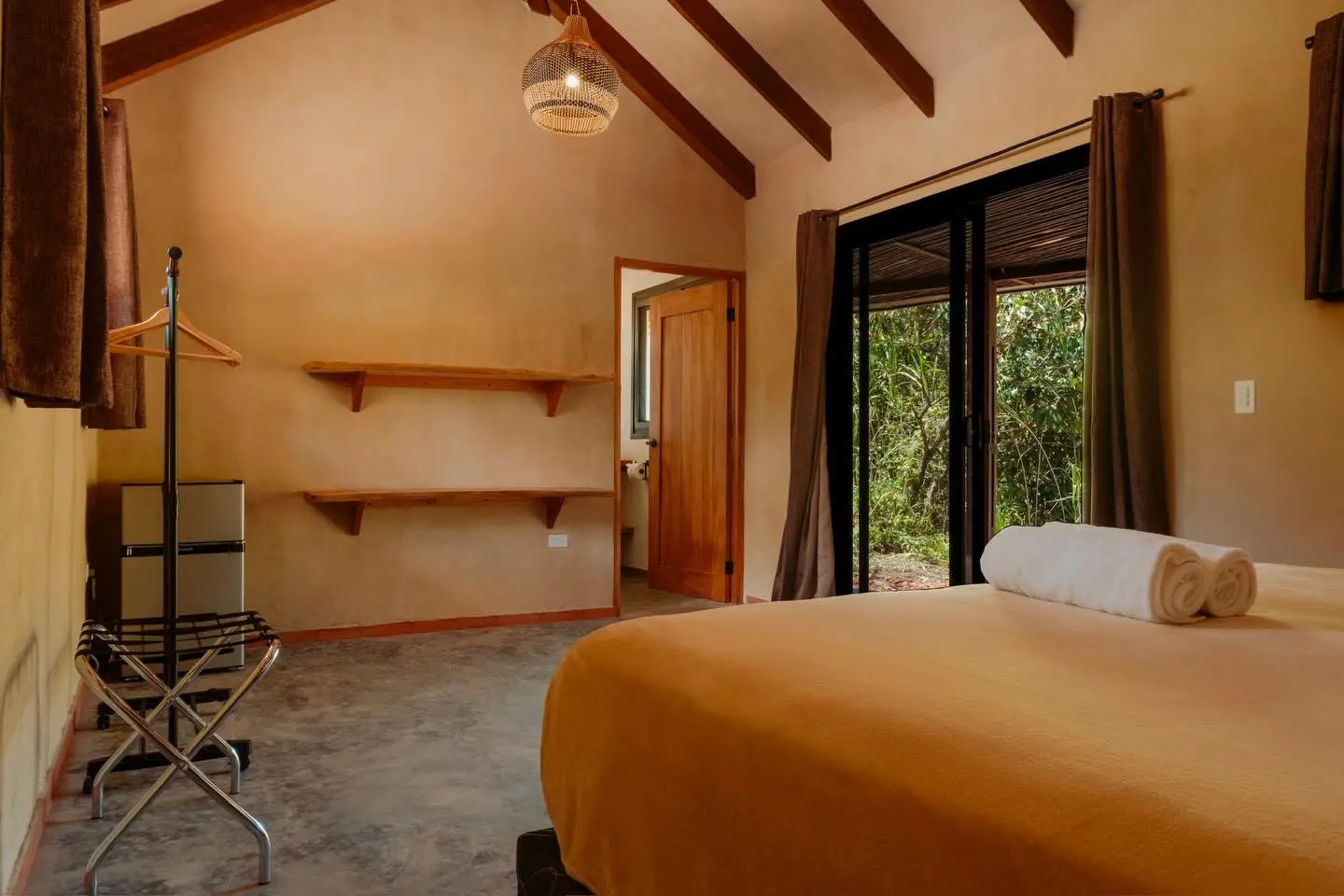 Habitación con puerta corrediza de vidrio hacia el verde, estantes de madera y cama con toallas enrolladas.