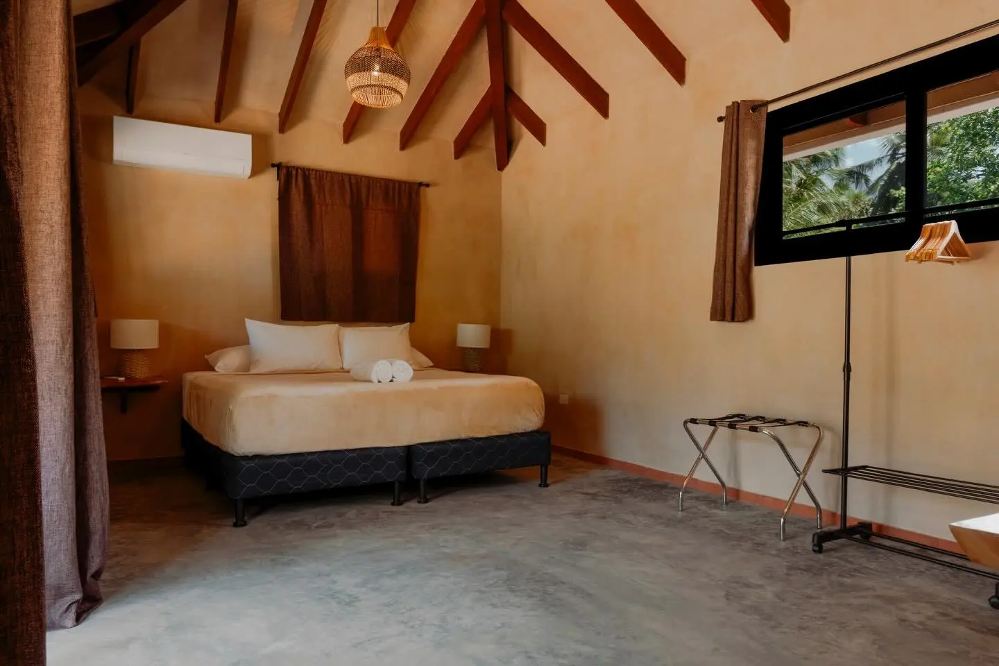 Habitación de techo alto con vigas de madera, lámpara tejida, cama con toallas y una ventana con vista a las palmeras.