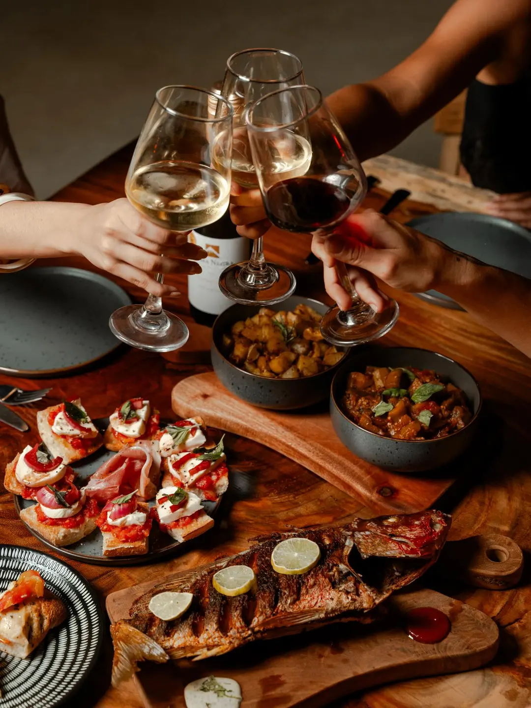 Brindis con copas de vino blanco y tinto sobre una mesa de madera con tostadas, papas, vegetales y un pescado entero con limón.