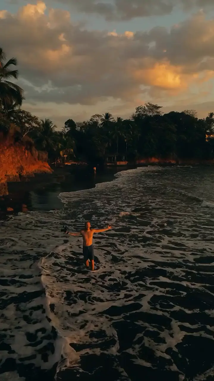 Persona de pie en una playa de arena oscura al atardecer, bajo nubes anaranjadas.