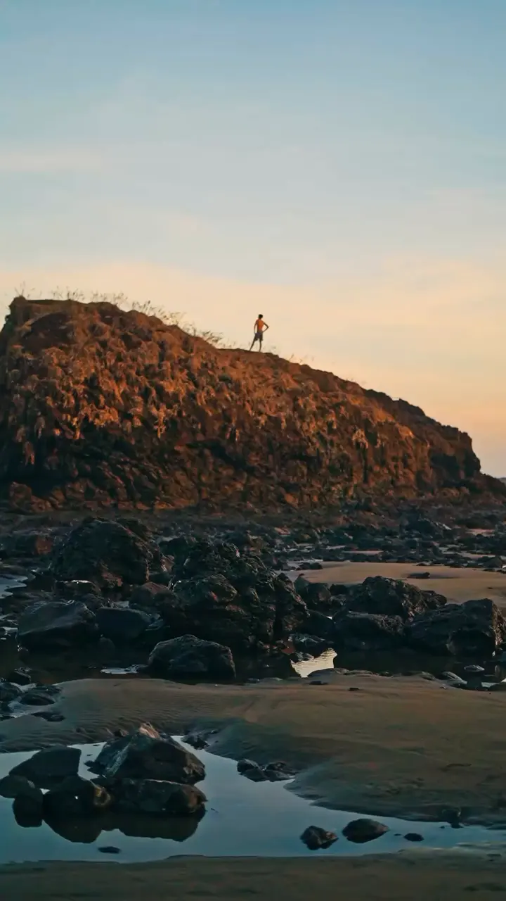 Islote rocoso sobre la playa al atardecer, con una persona de pie en lo alto.