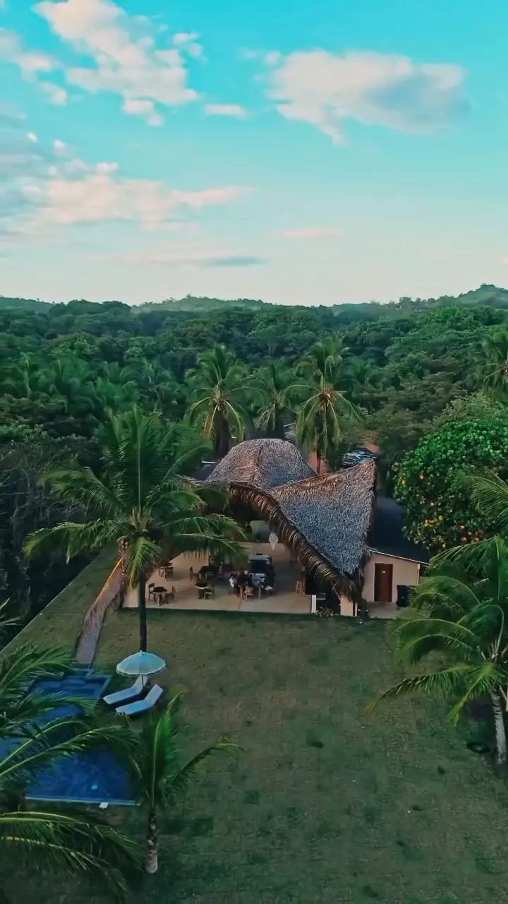 Vista aérea de una construcción de techo de palma entre selva y colinas.